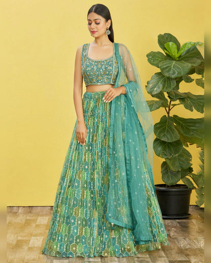 Woman in a green and blue patterned outfit standing against a yellow wall with a plant.