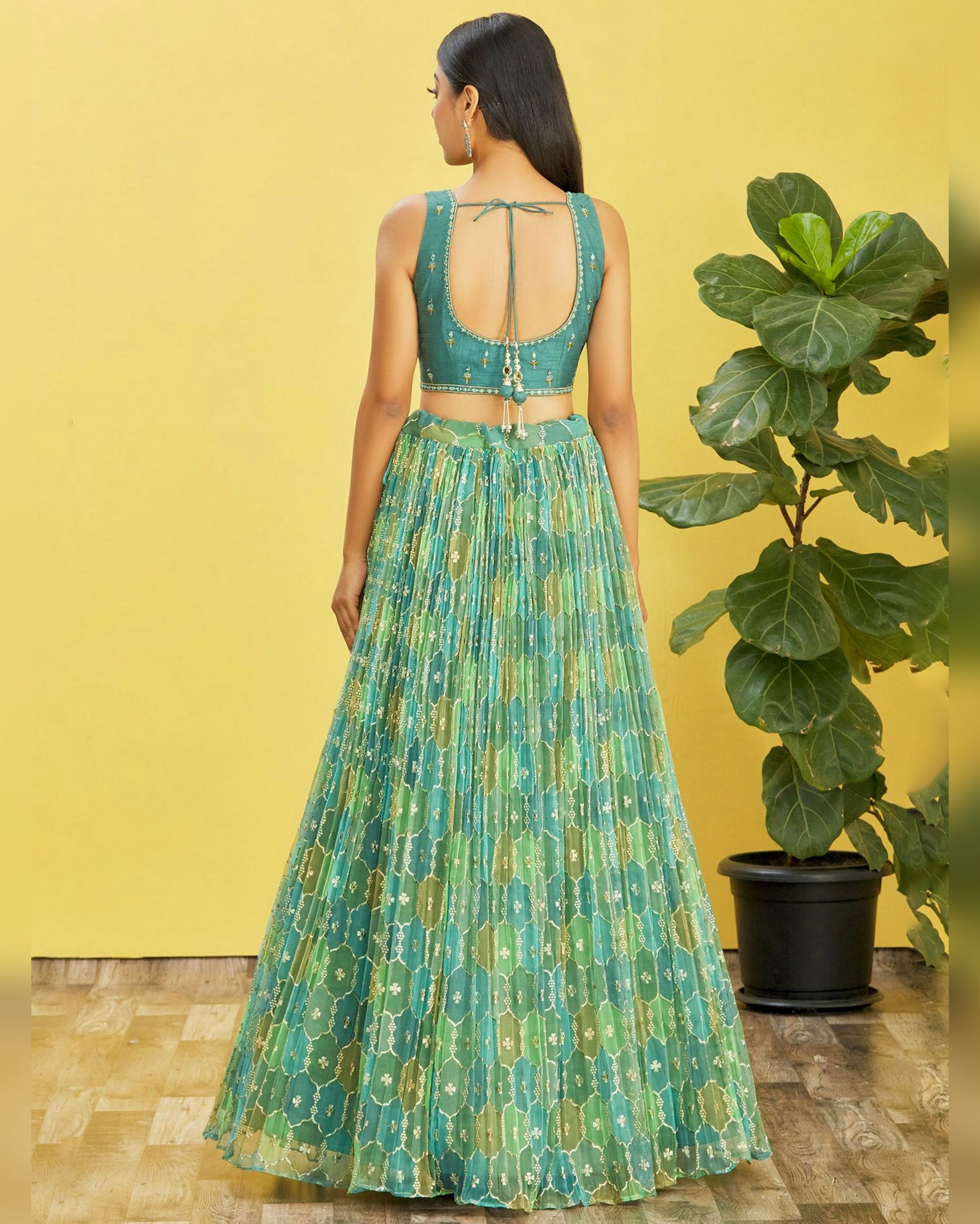 Woman wearing a green patterned outfit standing against a yellow wall with a plant.