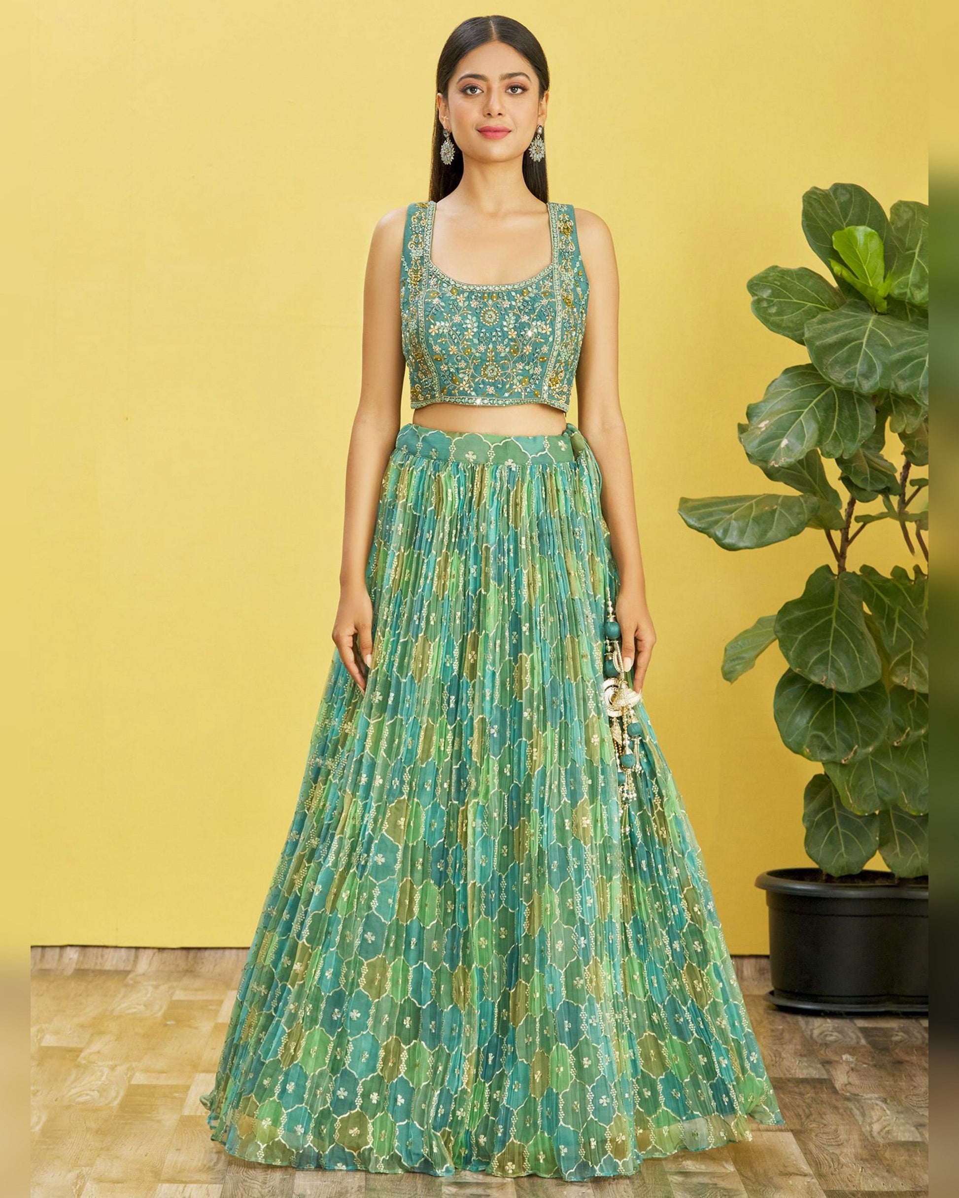 Woman wearing a green patterned outfit against a yellow wall with a plant.