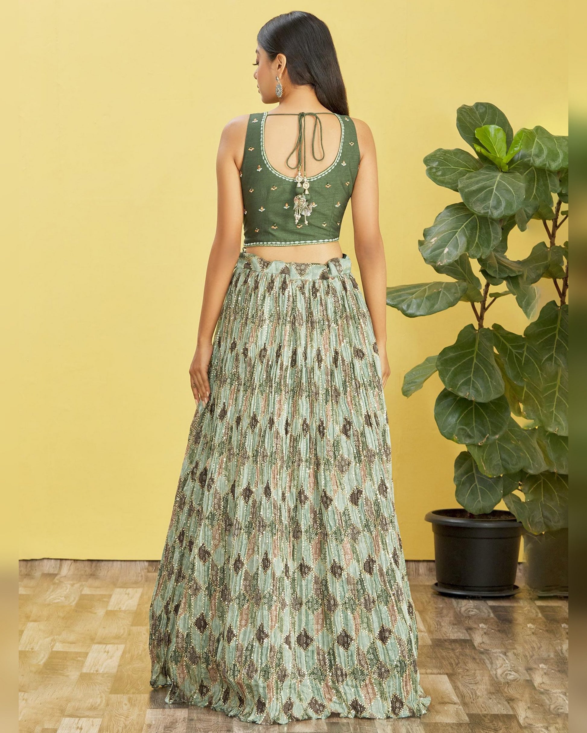 Woman wearing a green patterned outfit standing against a yellow wall with a plant in the foreground.