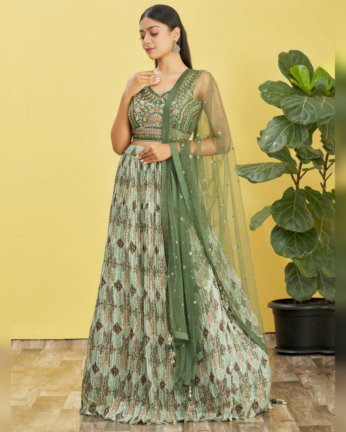 Woman in a green and patterned dress standing against a yellow wall with a plant.