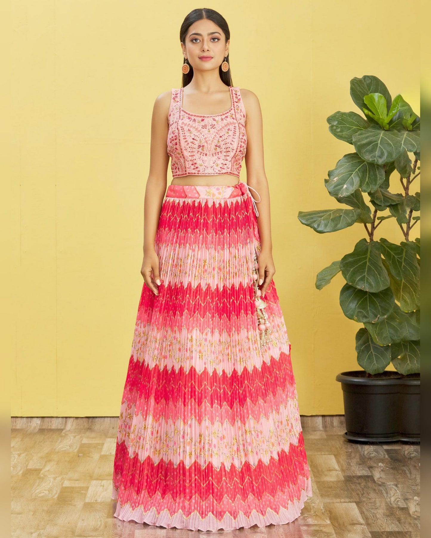 Woman wearing a pink and white patterned dress standing against a yellow wall with a plant in the background.