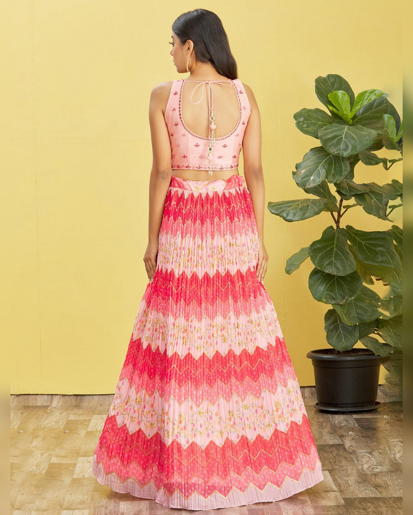 Woman wearing a pink and white patterned dress standing against a yellow wall with a plant.