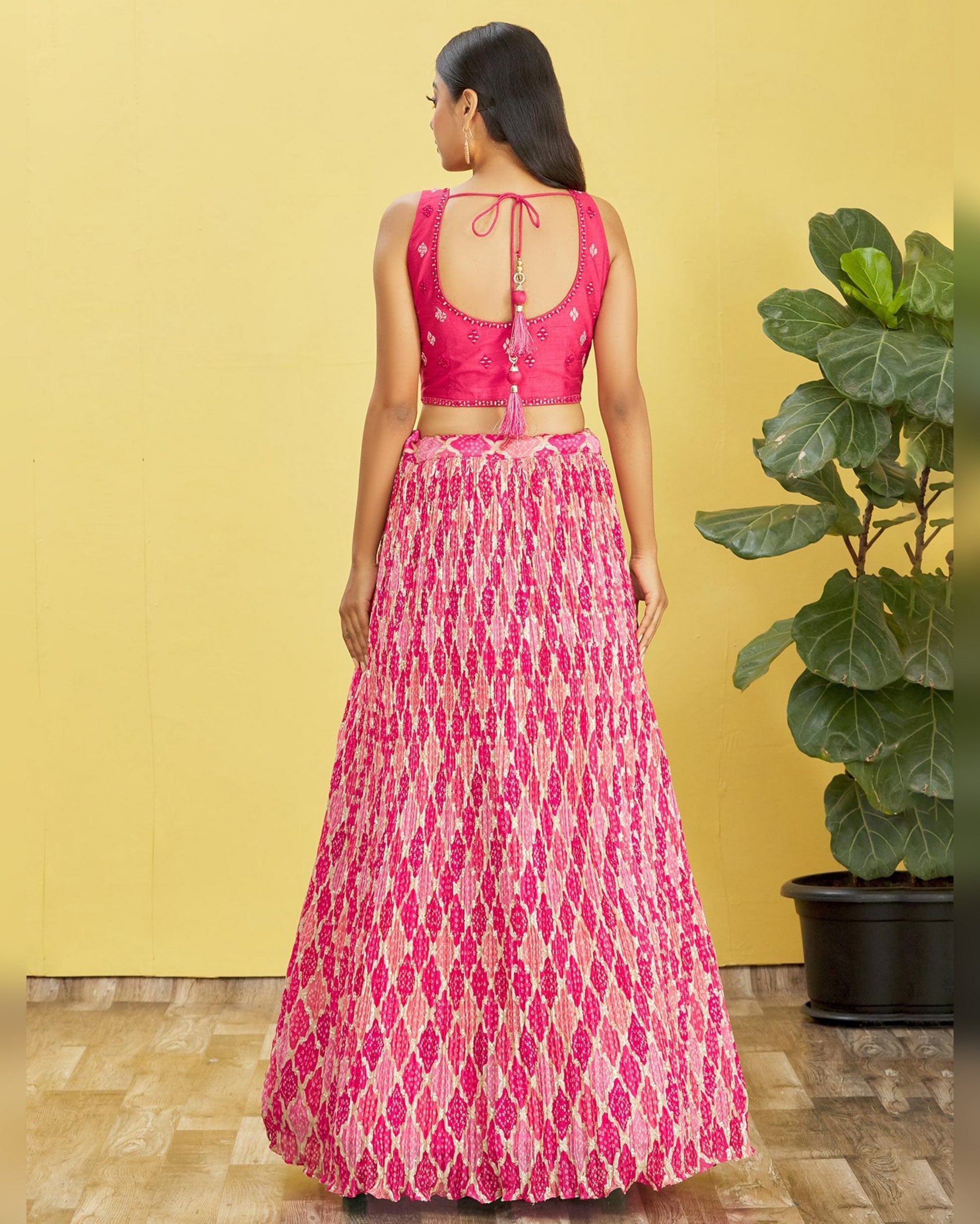 Woman wearing a pink and white patterned outfit standing against a yellow wall with a plant in the background.