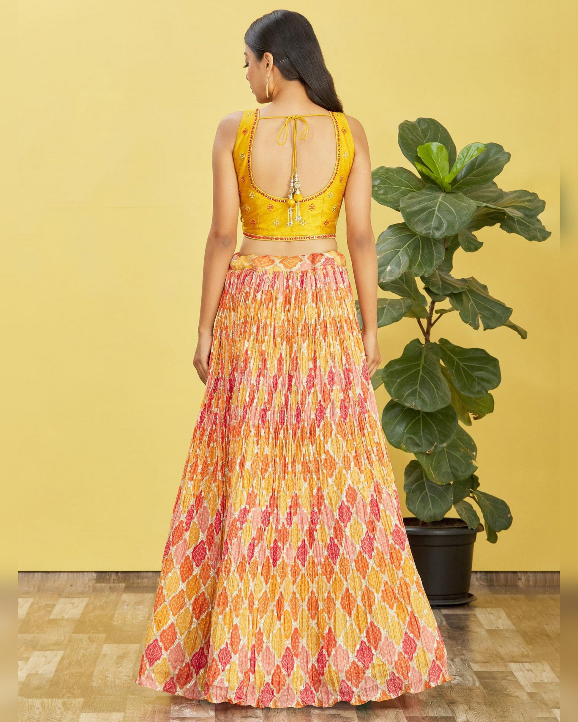 Woman wearing a yellow top and patterned skirt standing in front of a yellow wall with a plant.