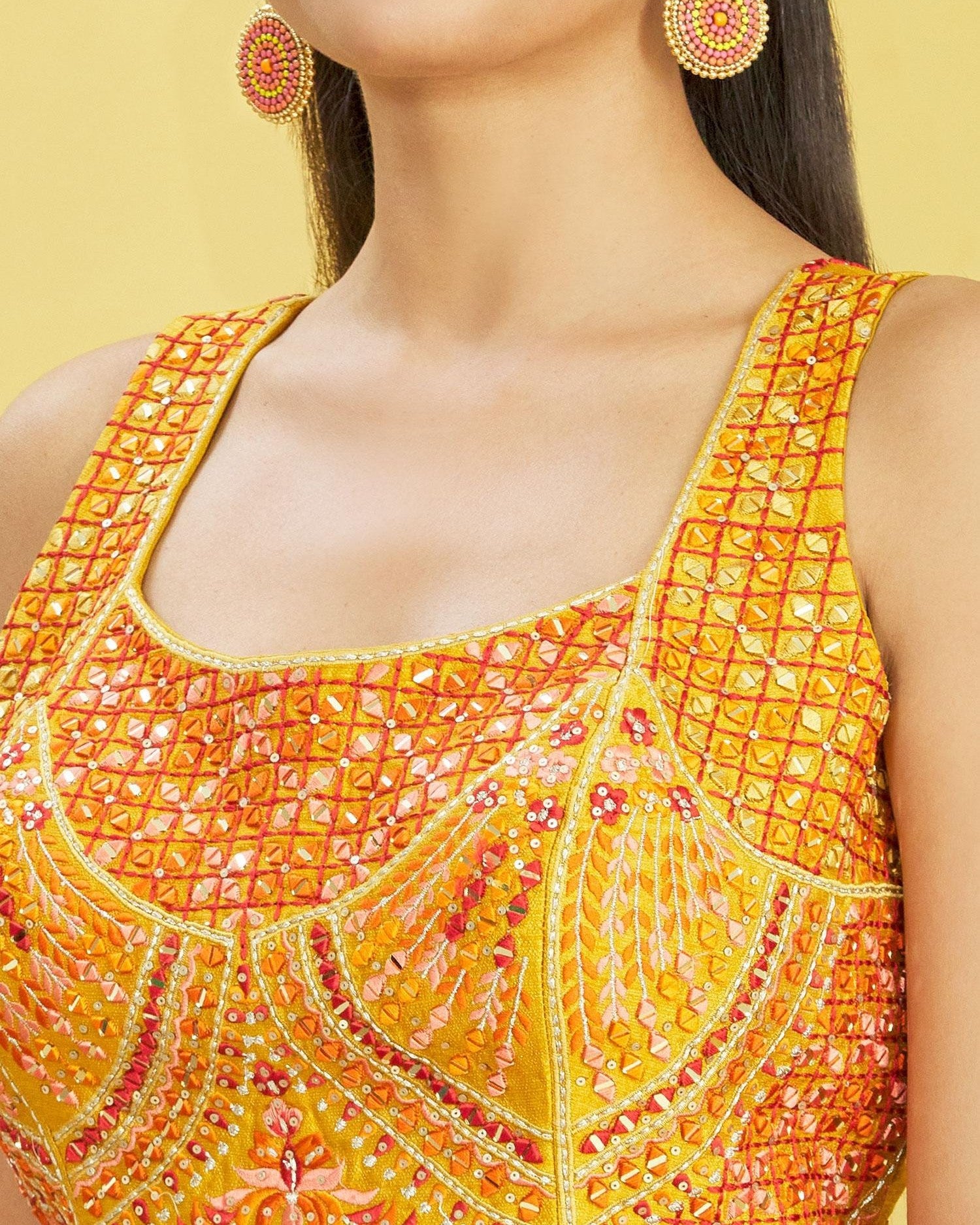 Close-up of a person wearing a yellow and orange embroidered top with matching earrings on a yellow background.