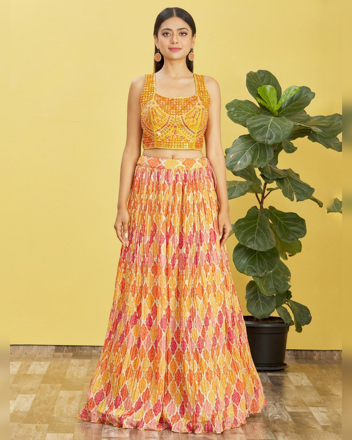 Woman wearing a yellow and orange patterned outfit standing next to a potted plant against a yellow wall.