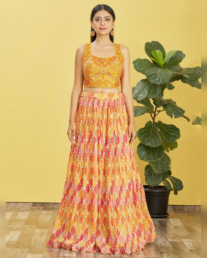 Woman wearing a yellow and orange patterned outfit standing next to a potted plant against a yellow wall.
