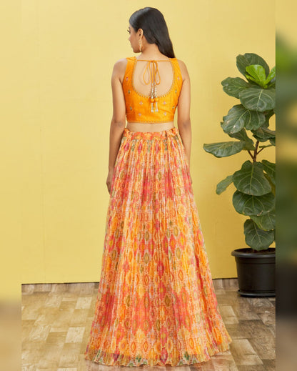 Woman wearing a bright orange top and patterned skirt against a yellow wall with a plant.