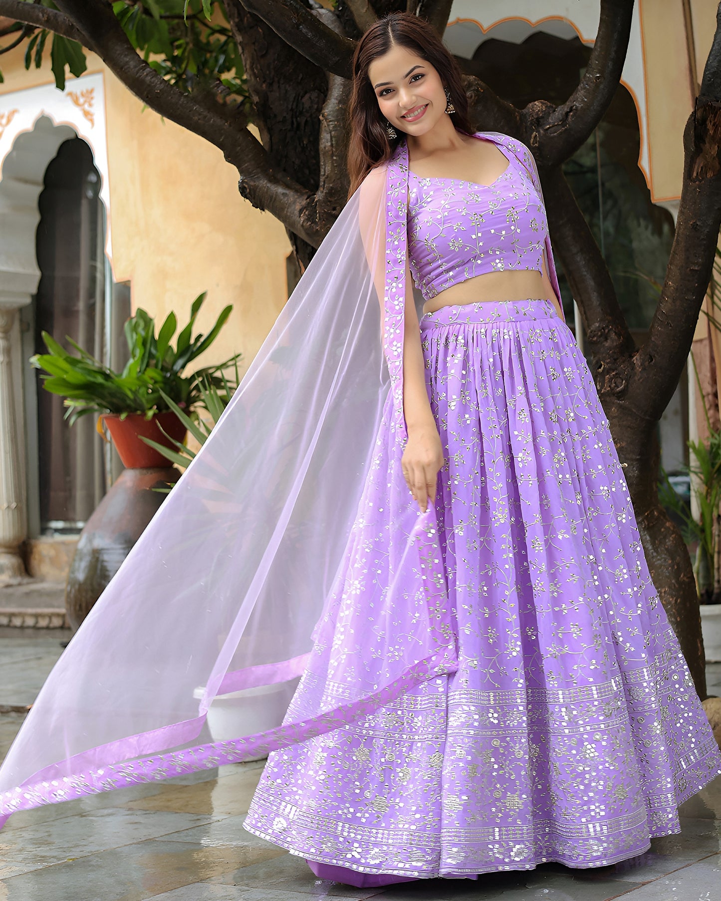 Woman in a purple traditional outfit standing outdoors with trees and plants in the background