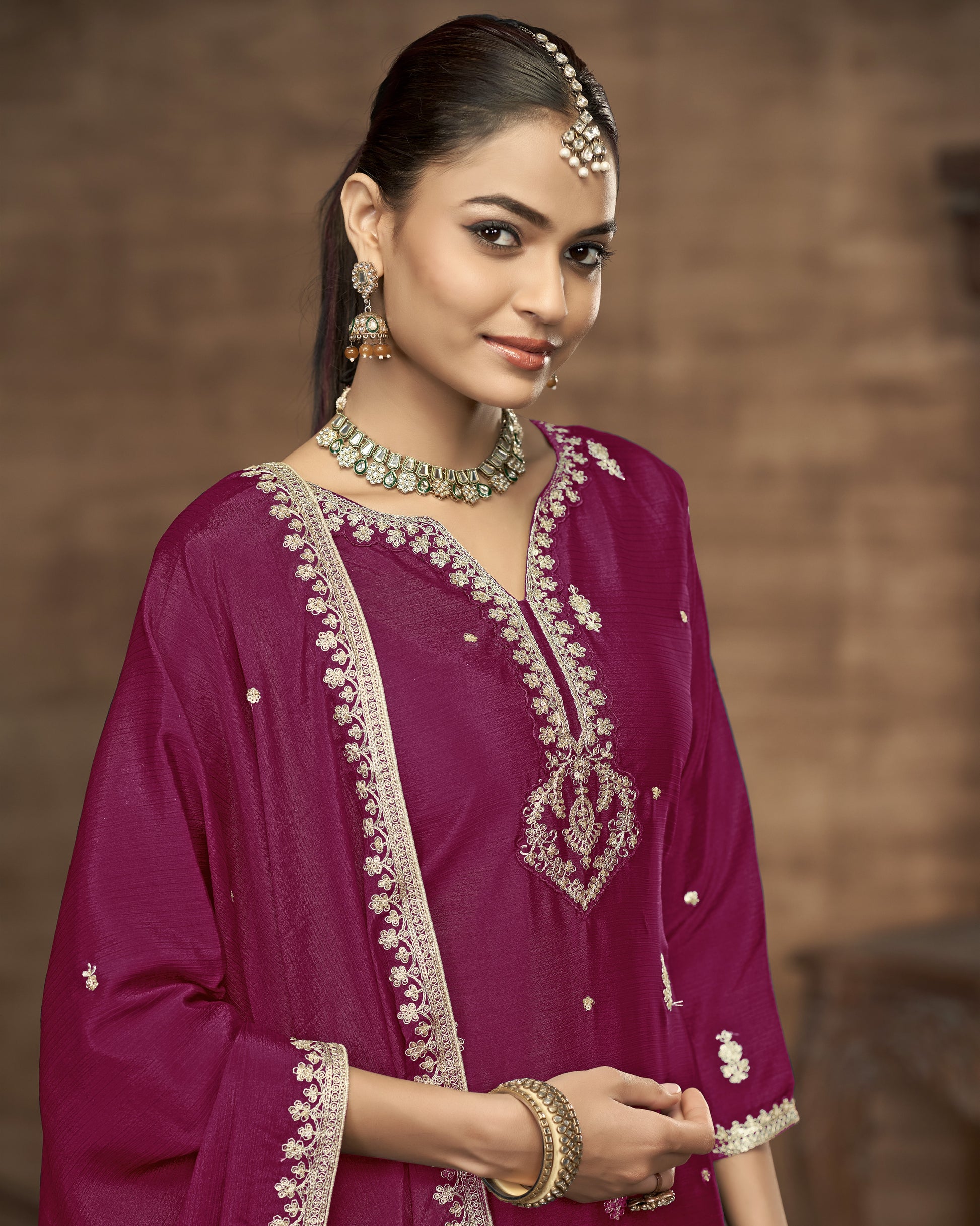 Woman wearing a traditional pink outfit with intricate embroidery and jewelry against a brown background