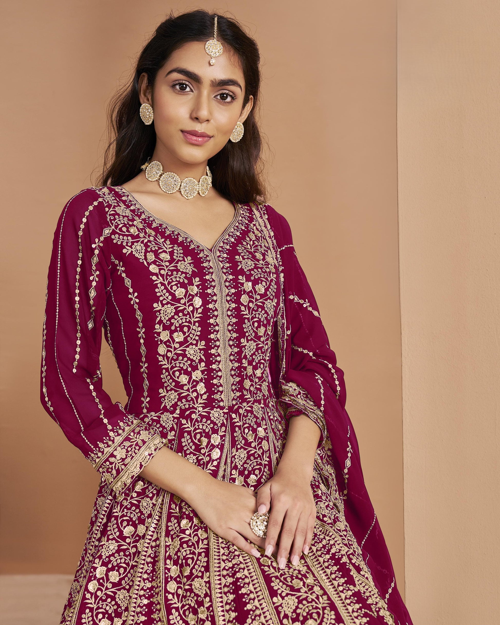 Woman wearing a maroon and gold traditional outfit with jewelry against a beige background