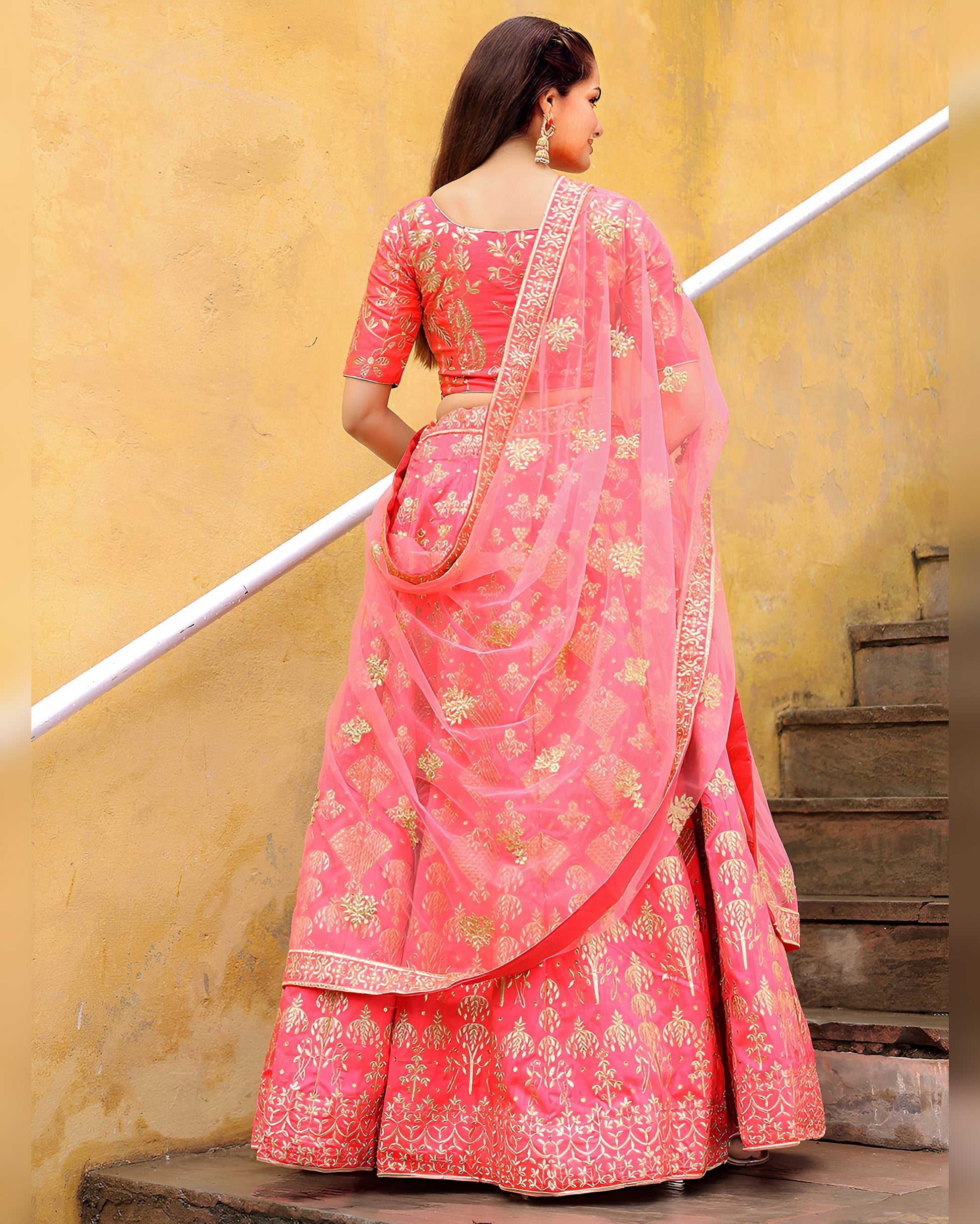 Woman wearing a pink saree with gold patterns against a yellow wall.