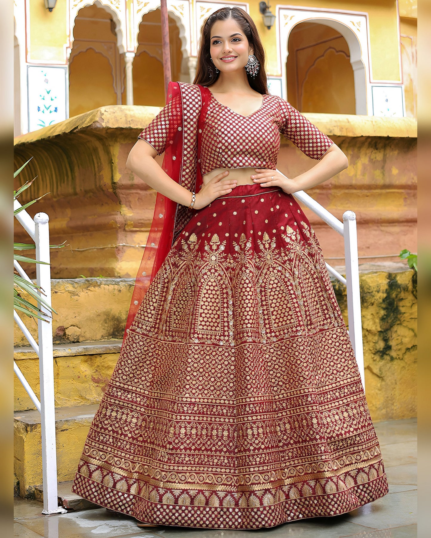 Woman in a traditional maroon and gold embroidered lehenga with a red dupatta against a yellow architectural background.