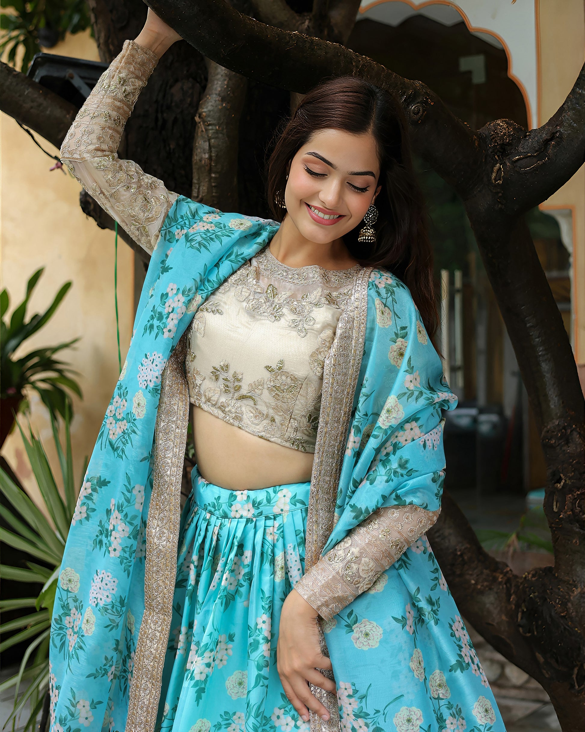 Woman in a blue floral saree with a beige blouse standing against a tree.