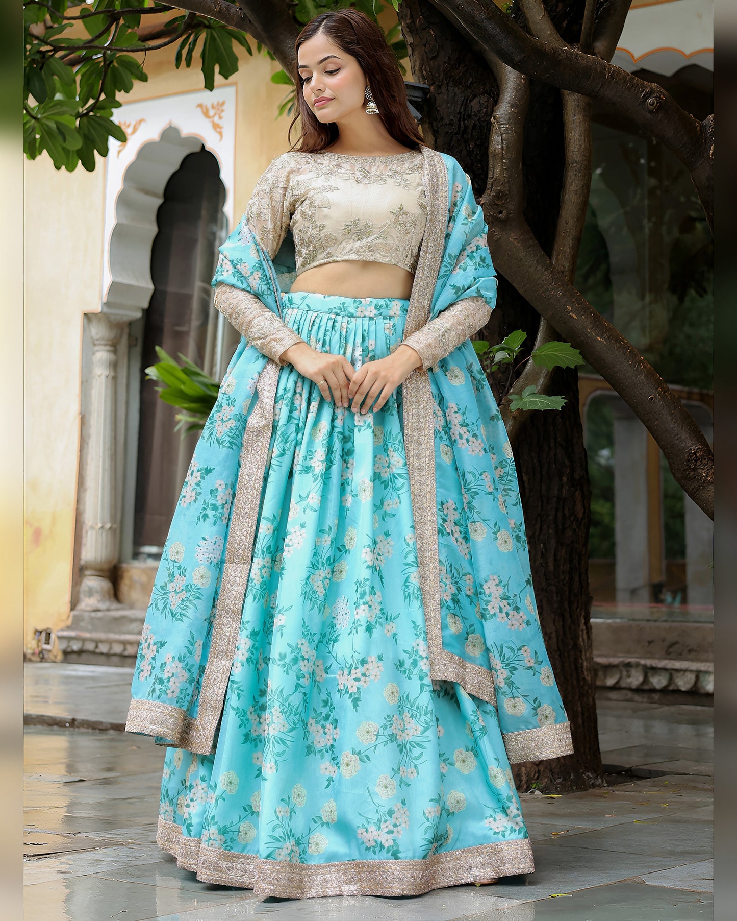 Woman in a light blue floral lehenga with beige blouse standing under a tree.