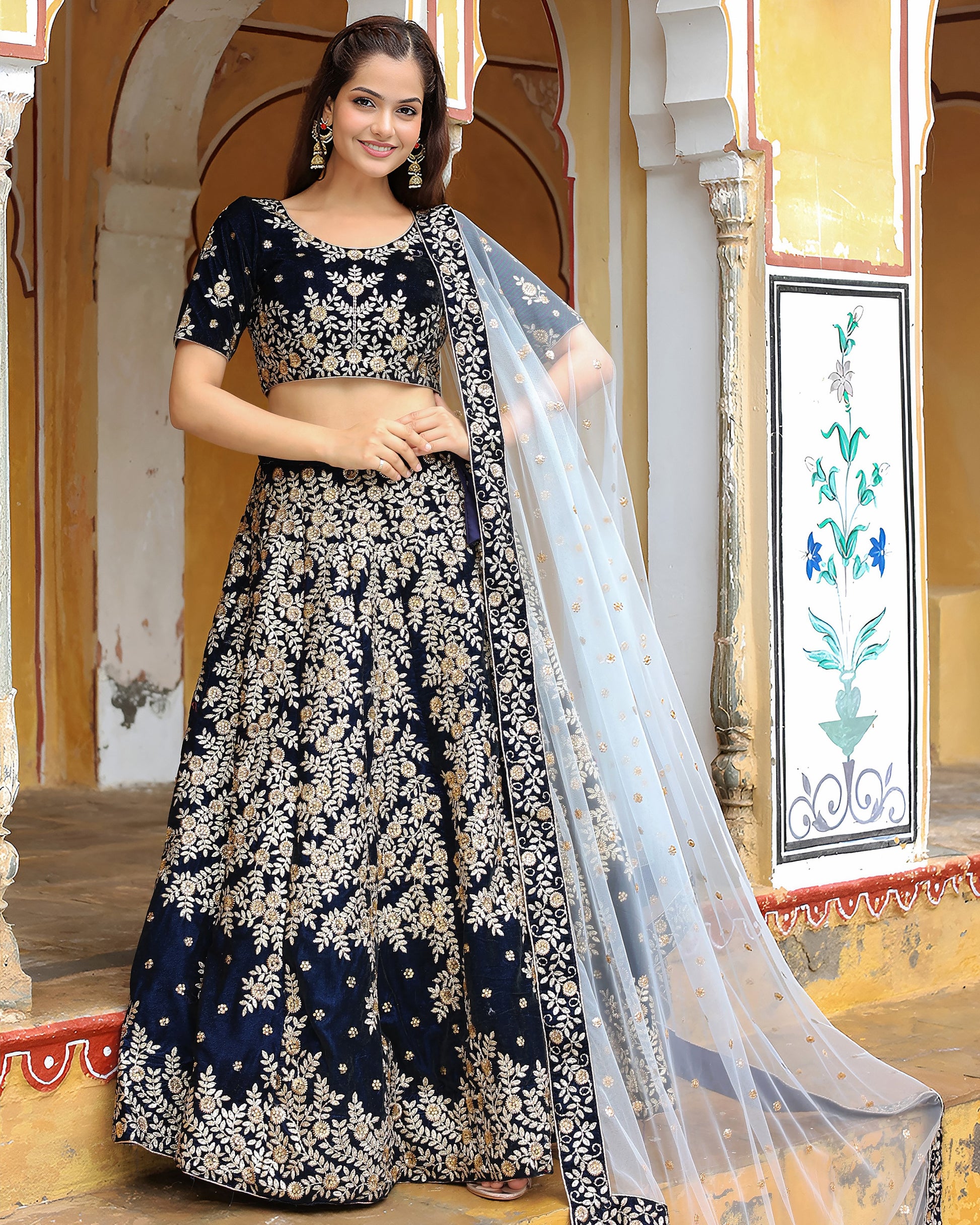 Woman wearing a navy blue and white embroidered traditional outfit with a sheer dupatta in an ornate indoor setting.