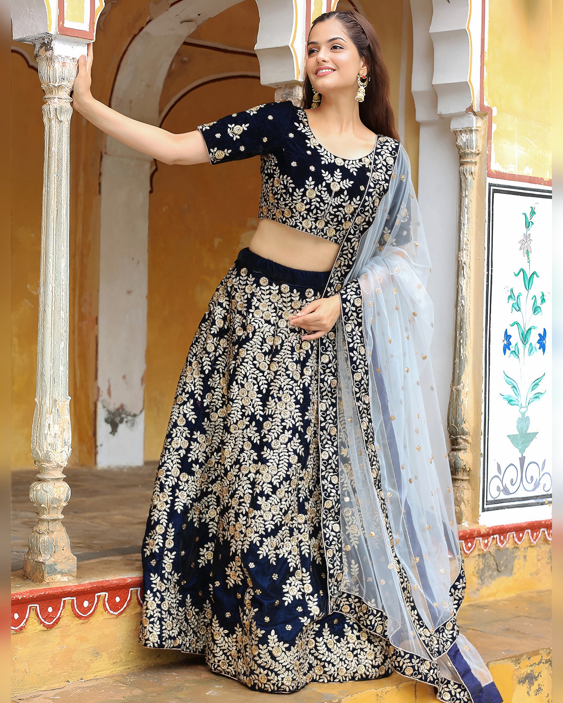 Woman in a traditional black and white floral outfit with a blue dupatta, standing in an ornate indoor setting.