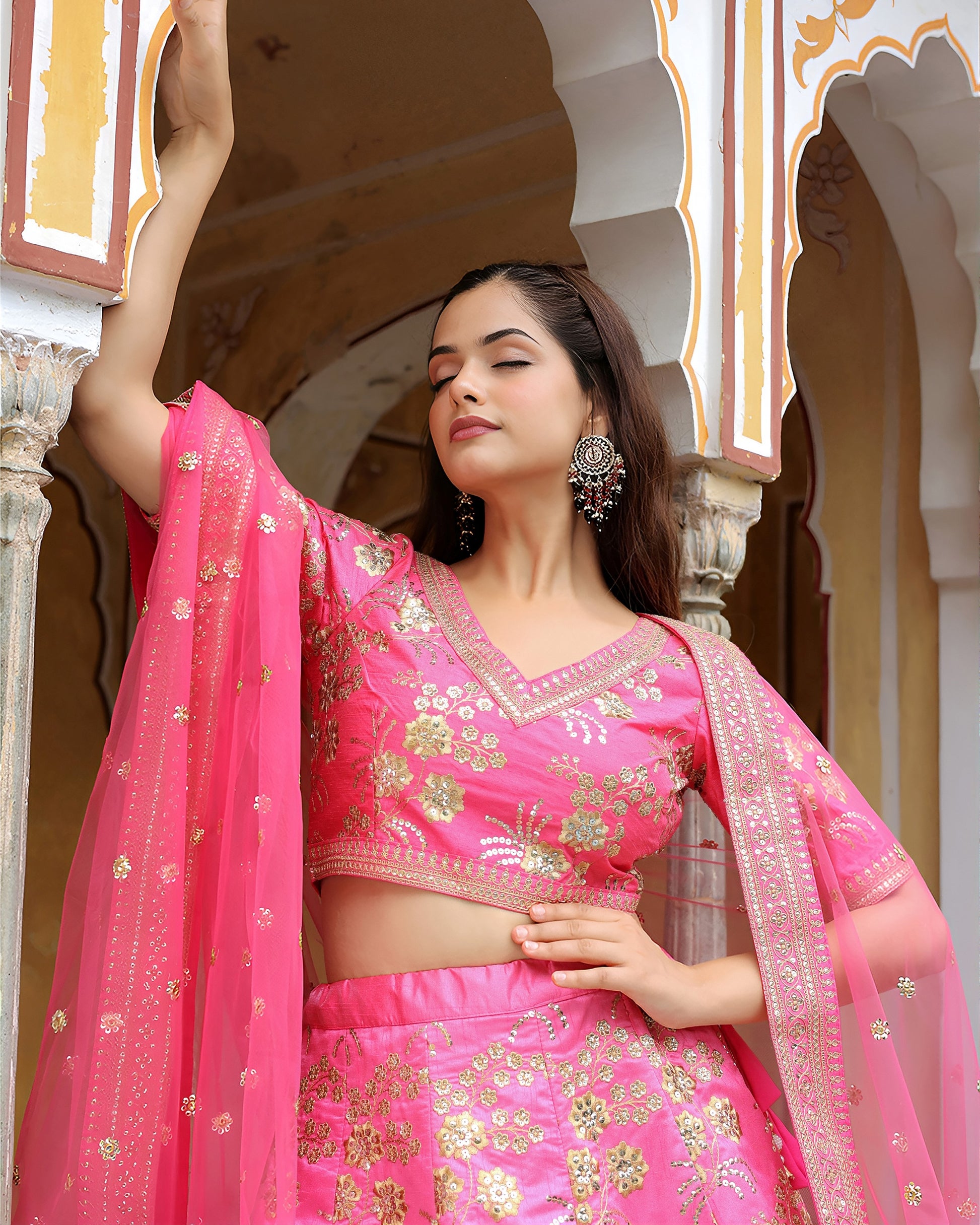 Woman in a pink traditional outfit with gold embroidery standing in front of an architectural background.