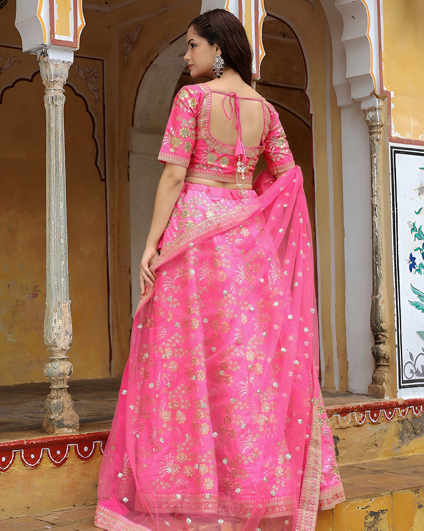 Woman in a pink saree with a floral pattern standing in an ornate architectural setting.