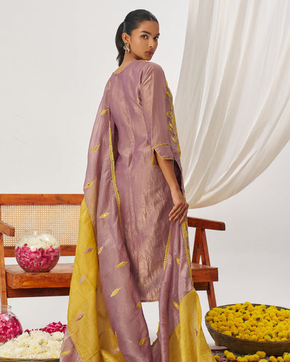 Woman in a traditional outfit with yellow and purple patterns standing in front of a white curtain.
