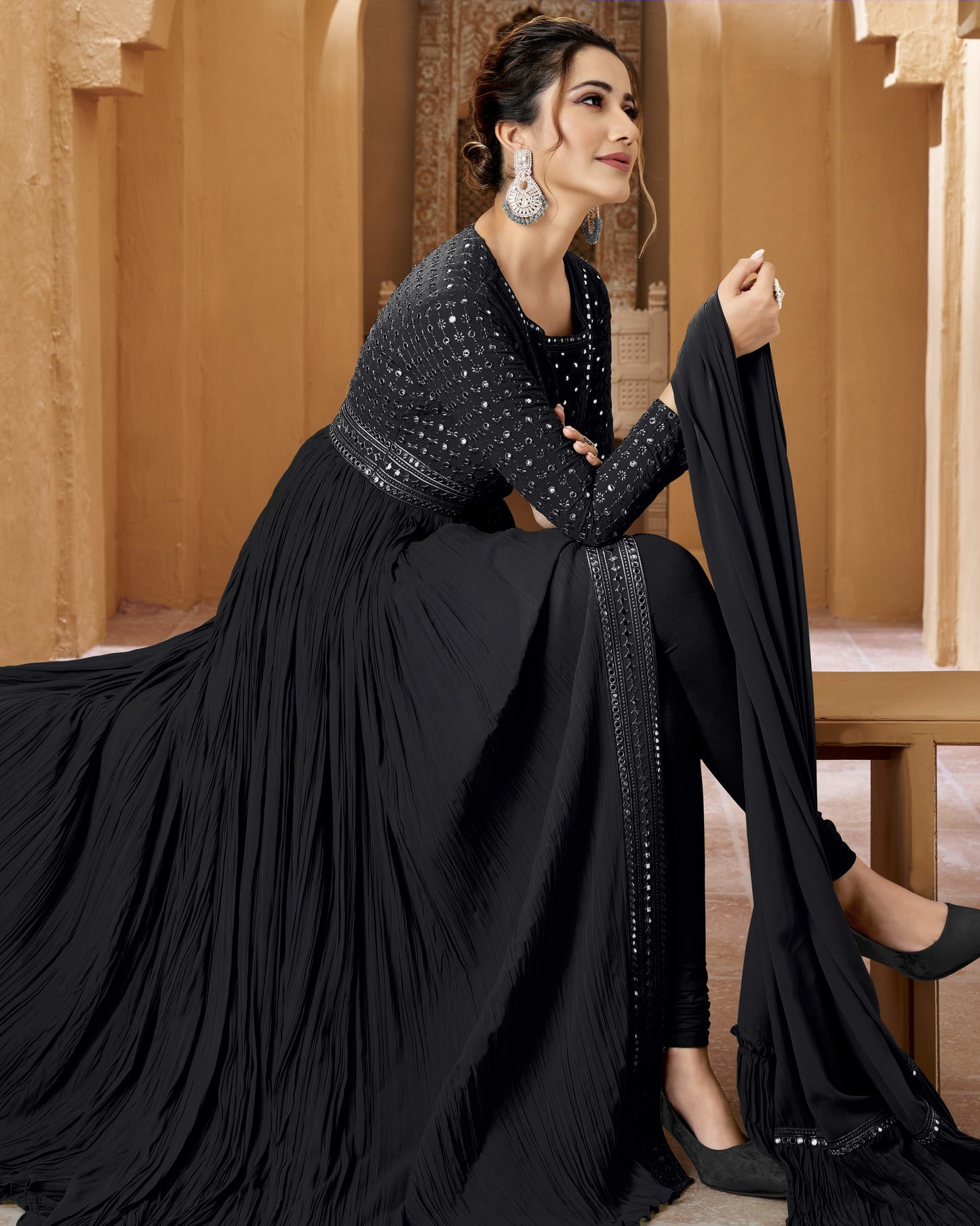 Woman in a black outfit with intricate detailing sitting in a warm-toned interior setting.