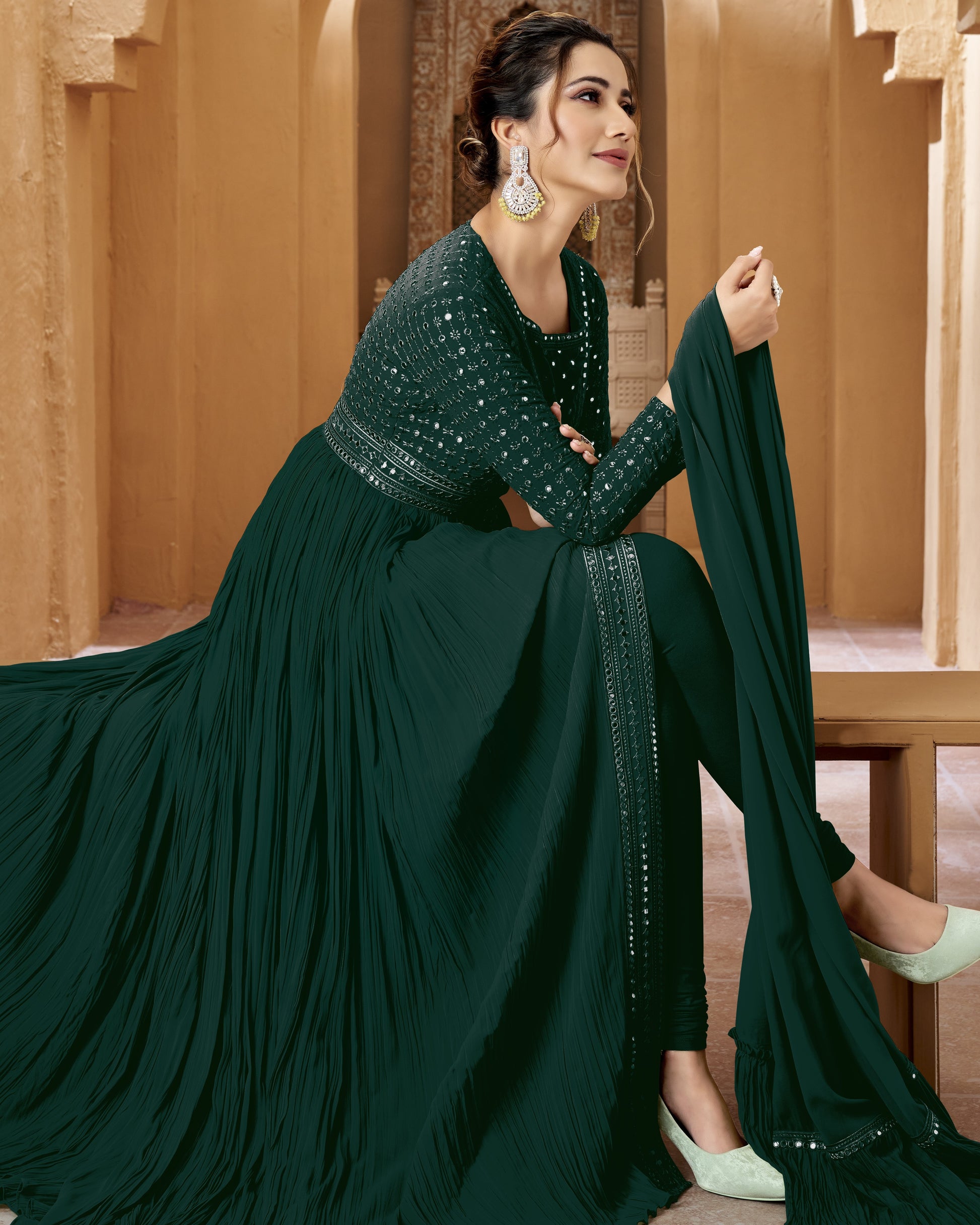 Woman in a dark green traditional outfit with embellishments, sitting against a warm-toned architectural background.