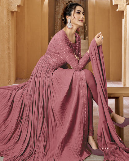 Woman in a pink traditional outfit with a flowing design, sitting against a beige wall.