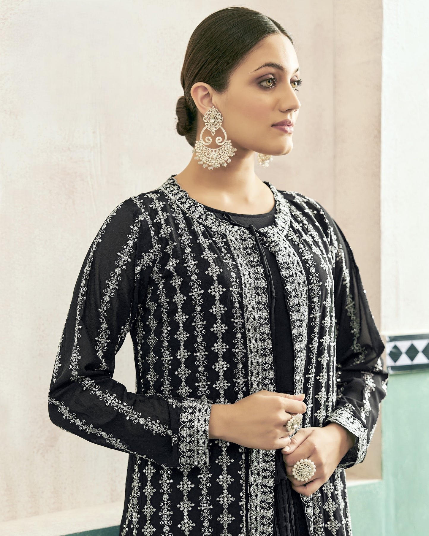 Woman wearing a black and silver embroidered outfit with earrings and rings indoors.