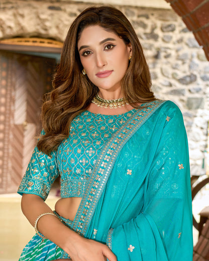 Woman wearing a teal saree with intricate patterns, standing against a stone wall background.