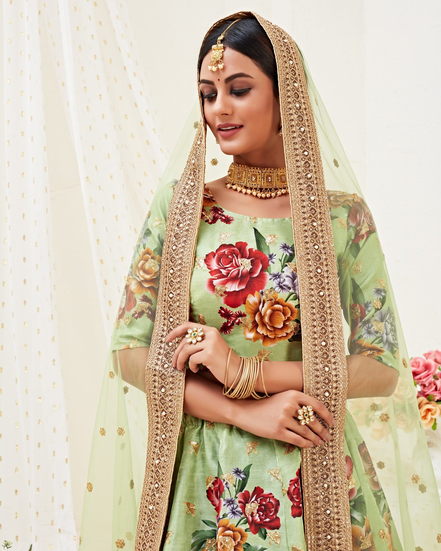 Woman in traditional green floral outfit with gold dupatta against a white curtain background