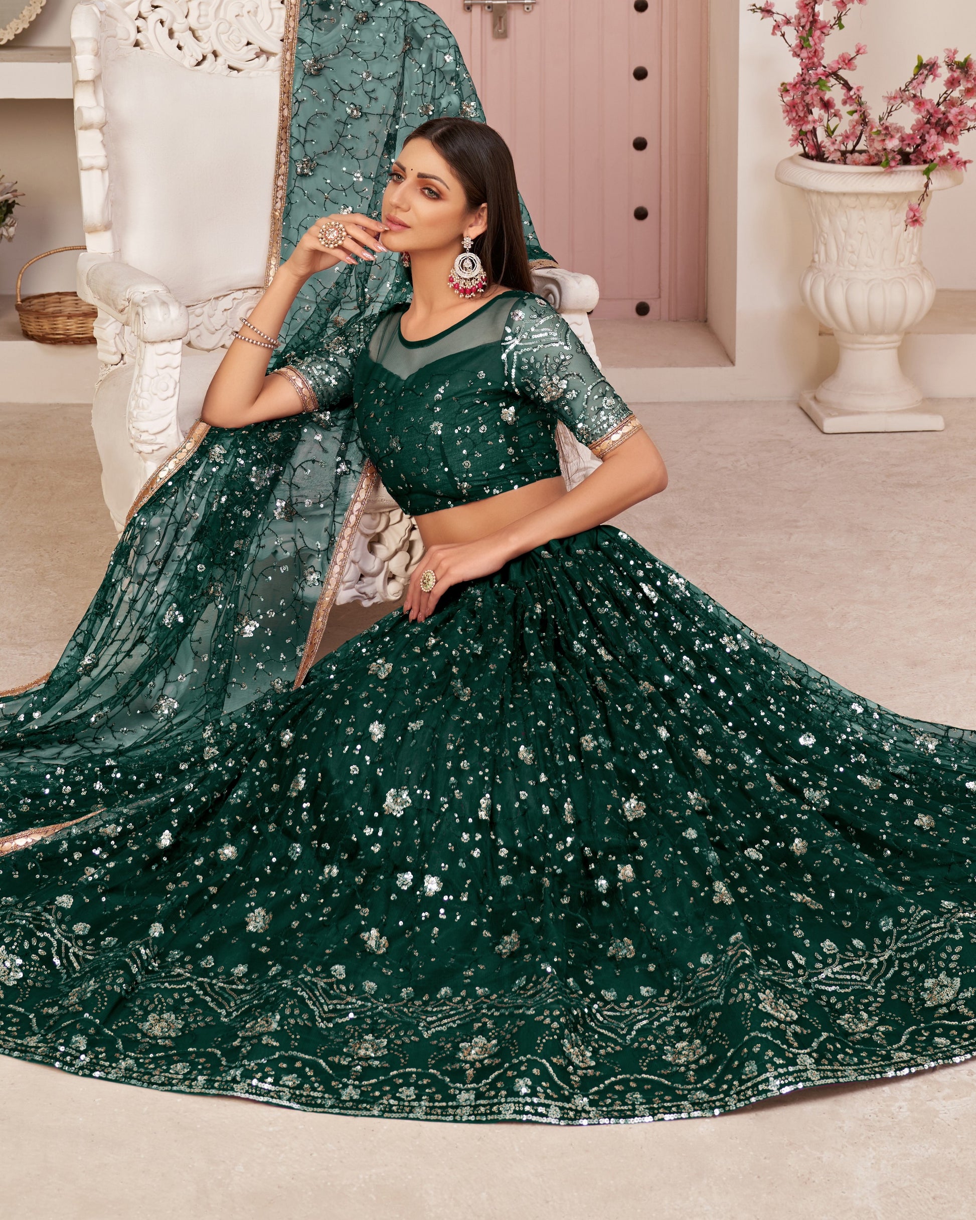 Woman in a green embroidered saree sitting on a white chair in a decorated room.