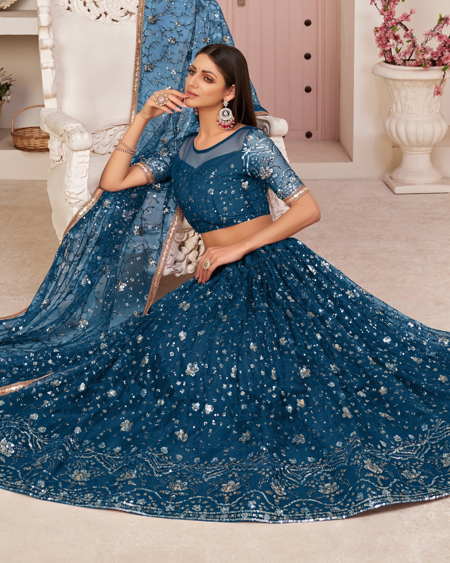 Woman in a blue embroidered saree sitting on a white chair in a decorated room.