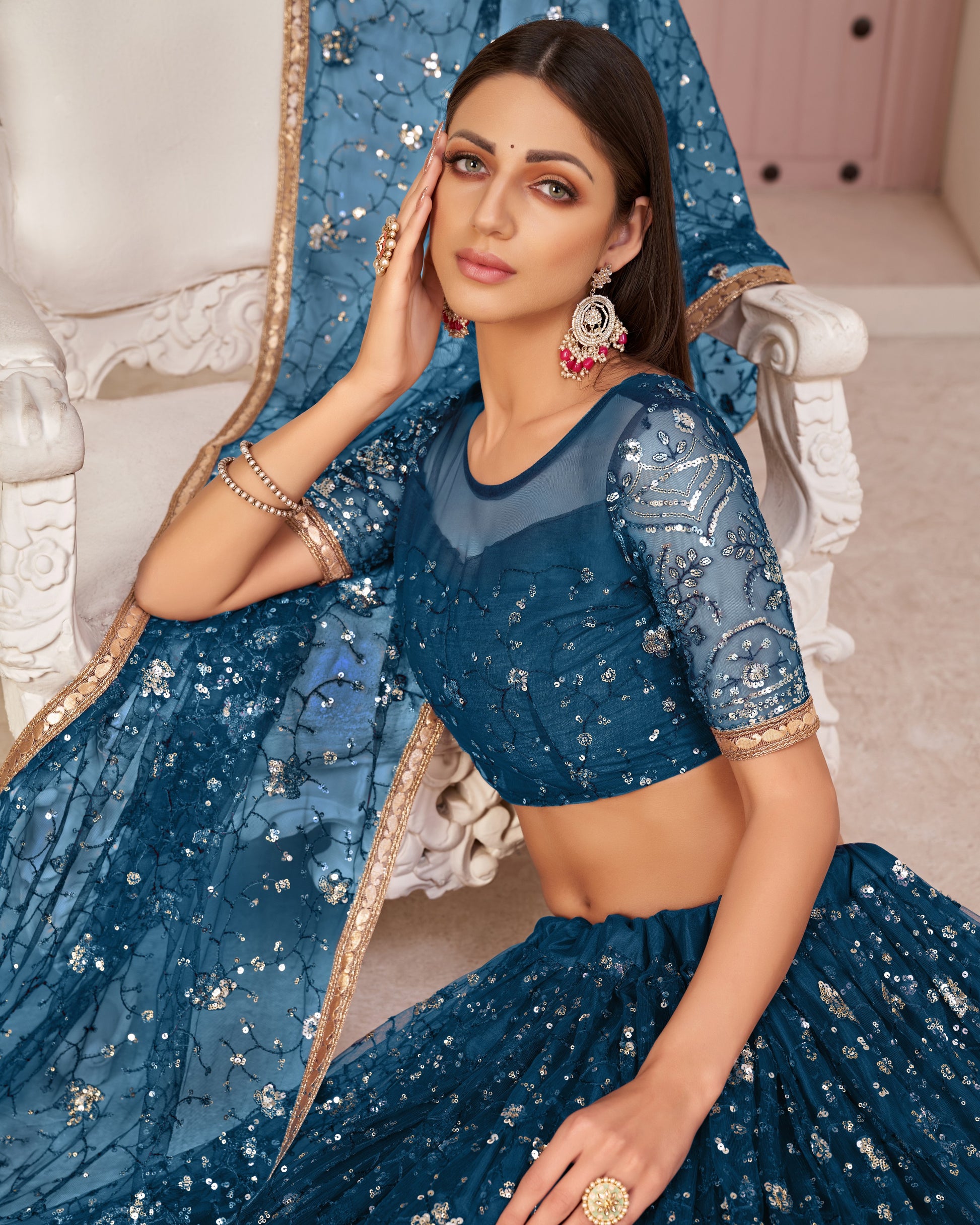 Woman in a blue embroidered traditional outfit sitting on a decorative chair.