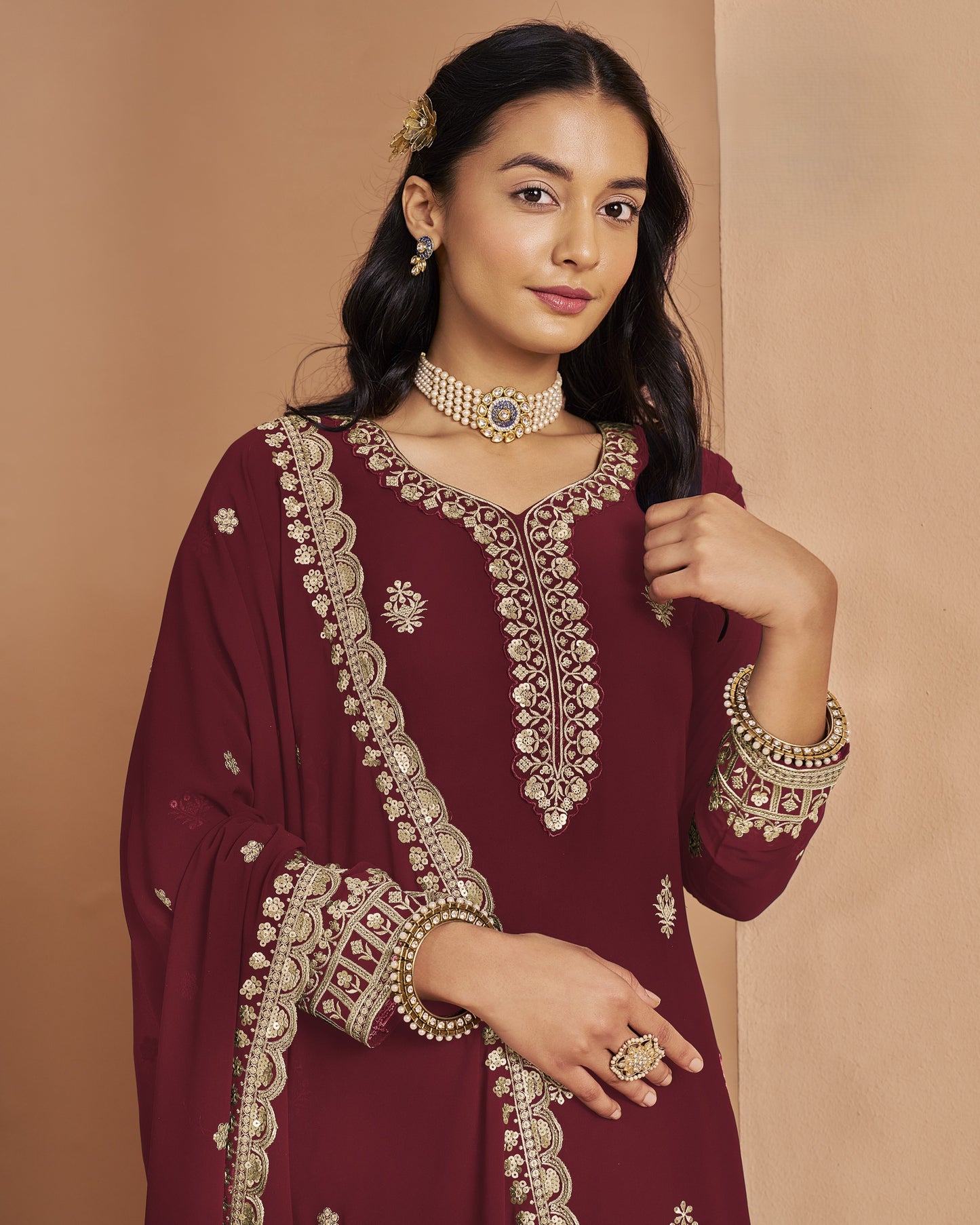 Woman wearing a maroon embroidered traditional outfit with gold jewelry against a beige background
