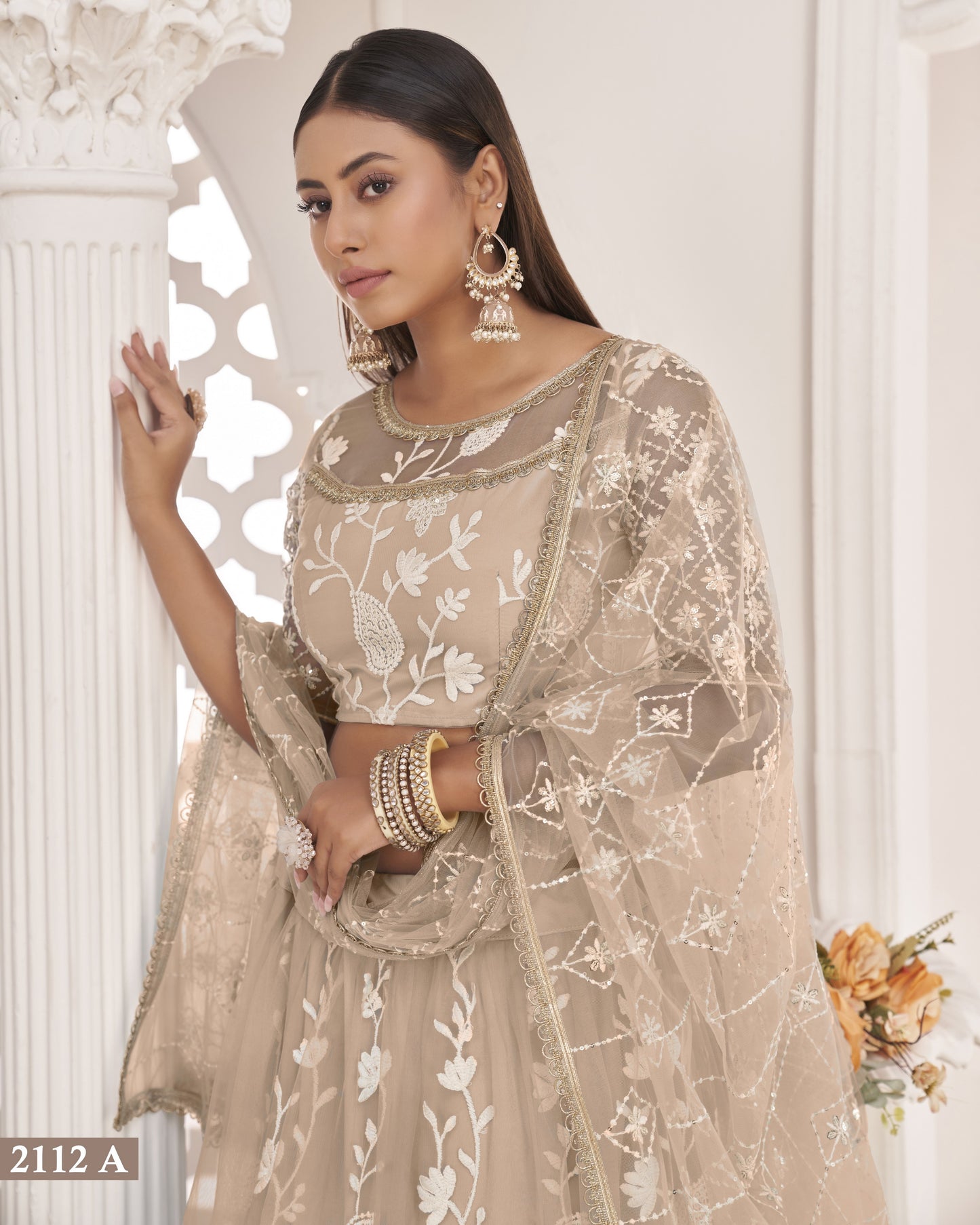 Woman wearing a beige embroidered traditional outfit with jewelry in a decorative setting.
