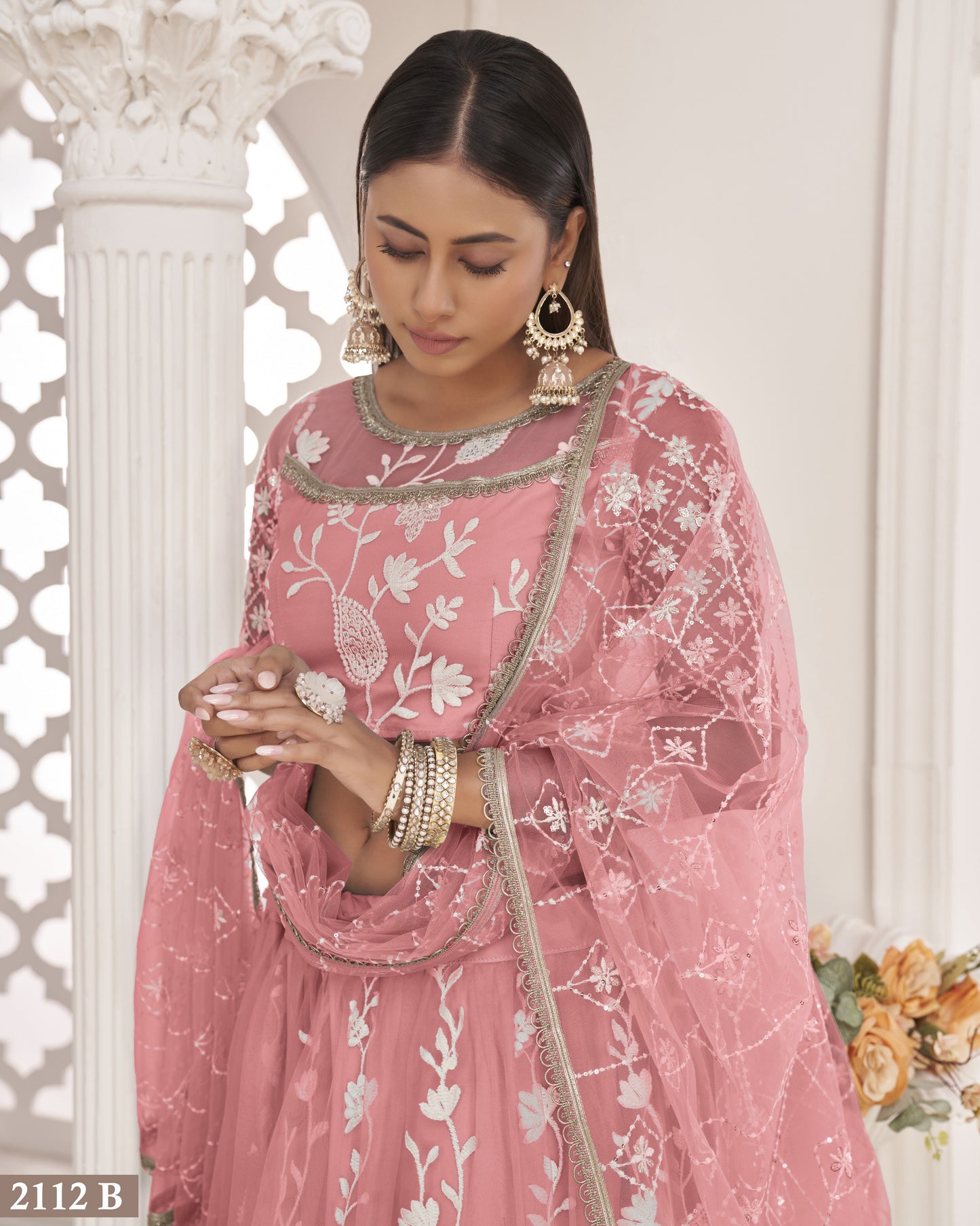 Woman wearing a pink embroidered traditional outfit with floral patterns, standing against a white decorative wall.