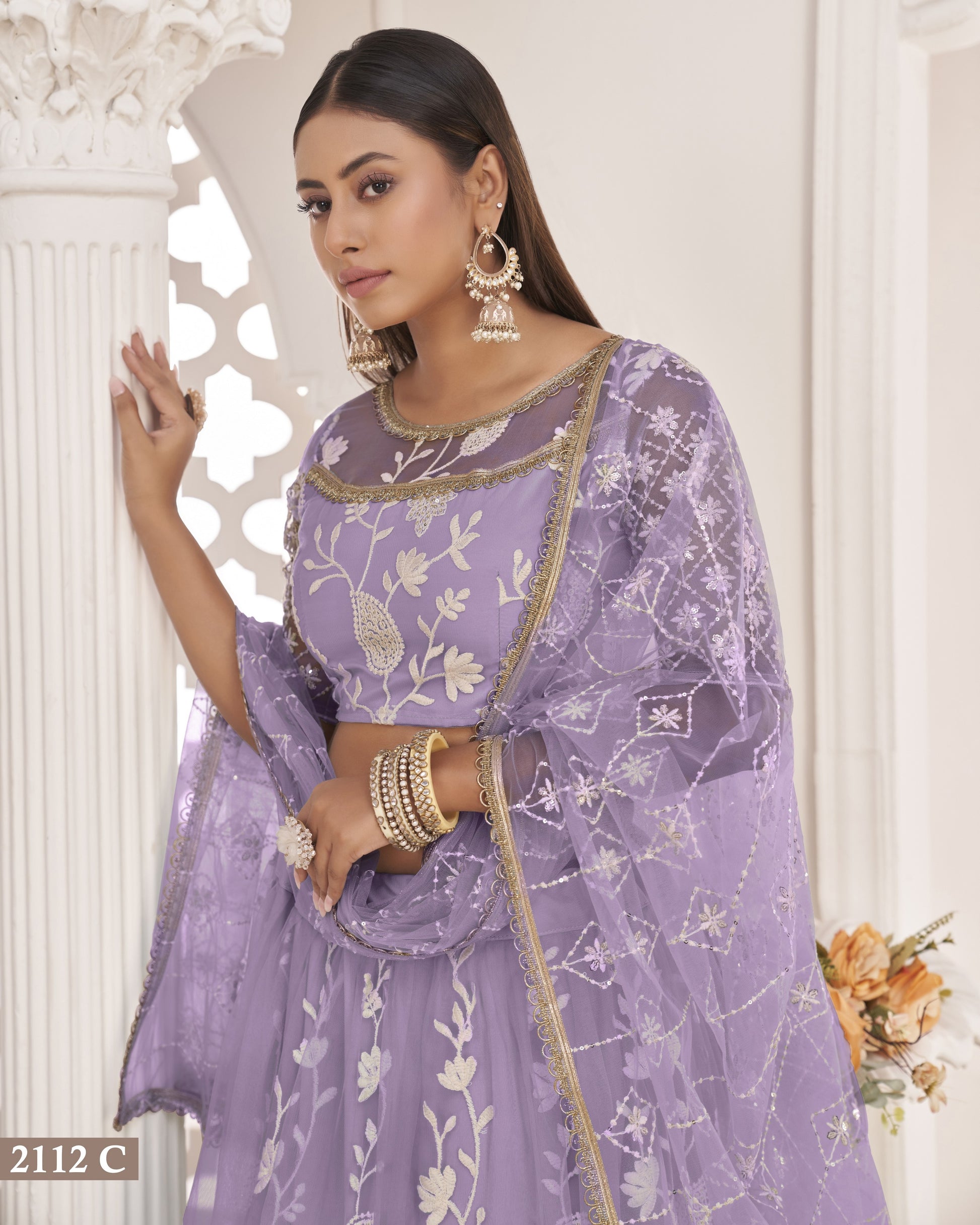 Woman wearing a purple embroidered traditional outfit with gold details against a white decorative wall.