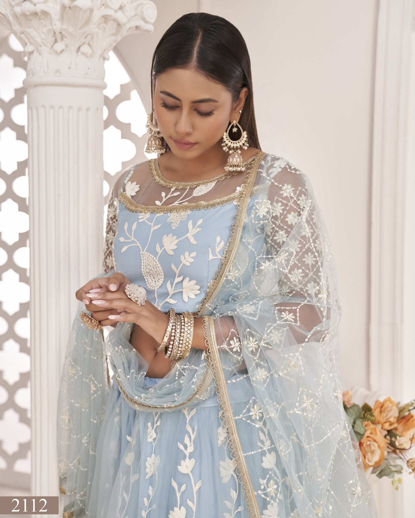 Woman in a light blue embroidered outfit with floral patterns, standing against a white wall.