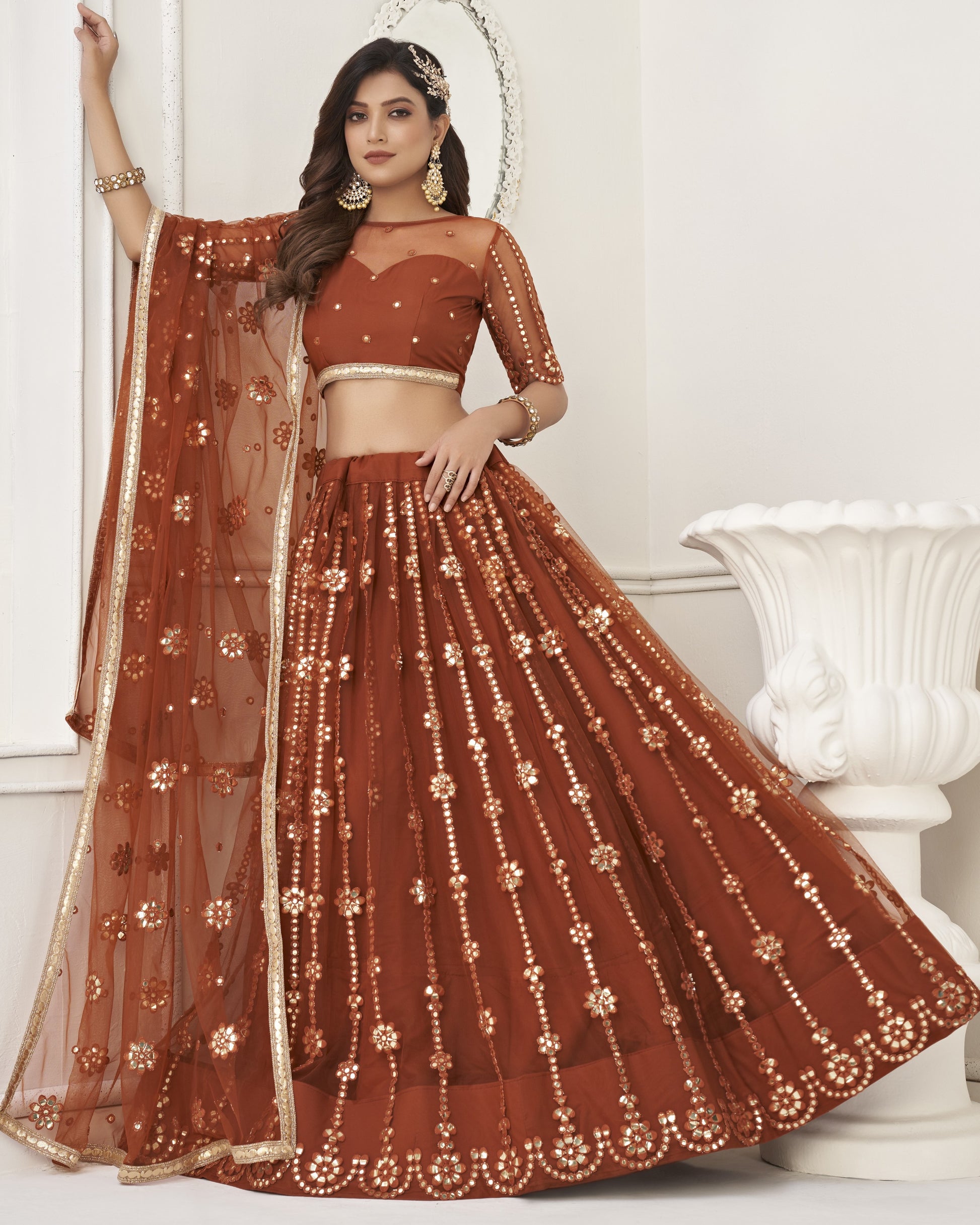 Woman wearing a rust-colored embroidered lehenga with a sheer dupatta in a white room.