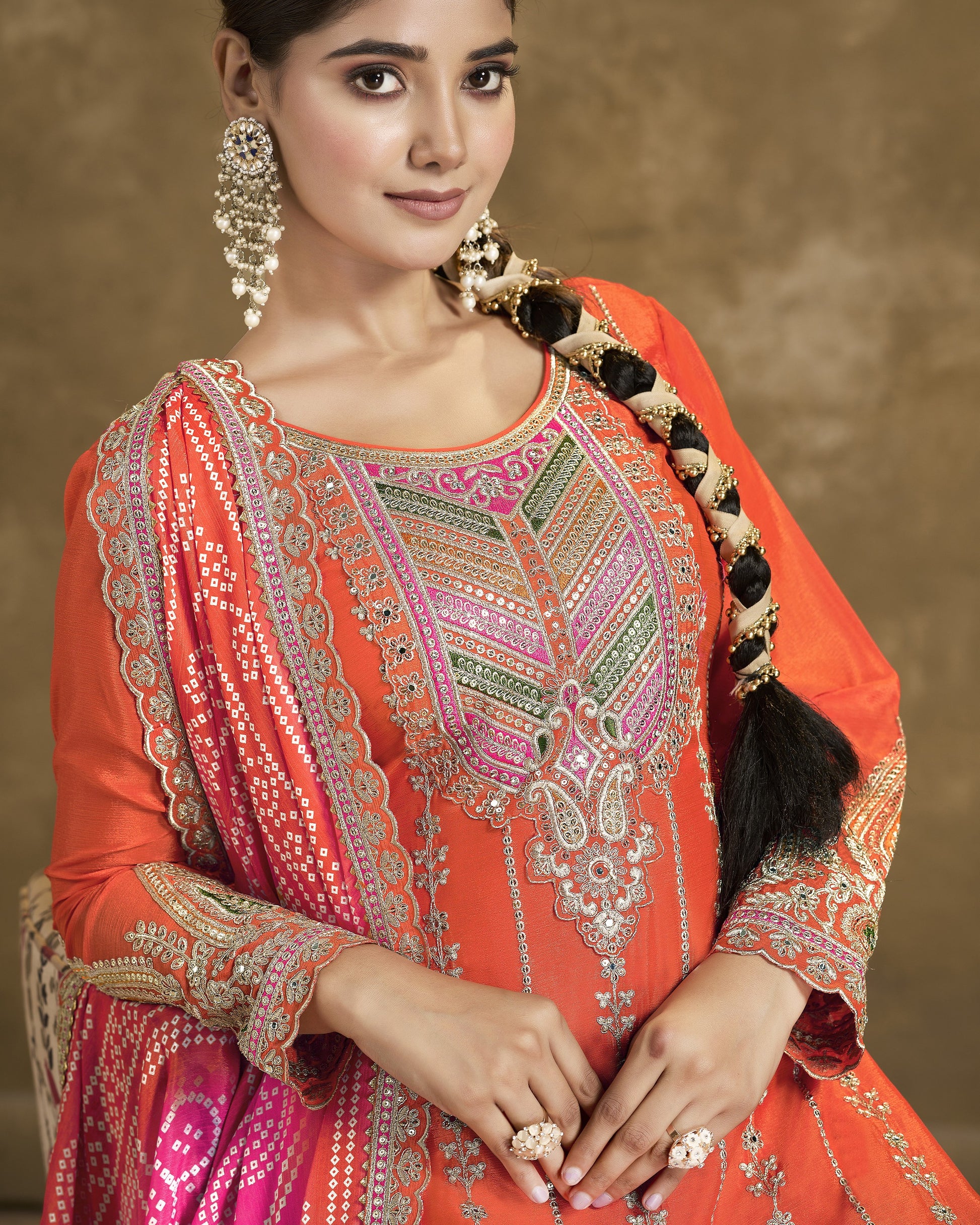 Woman wearing an orange and pink embroidered traditional outfit with jewelry against a brown background