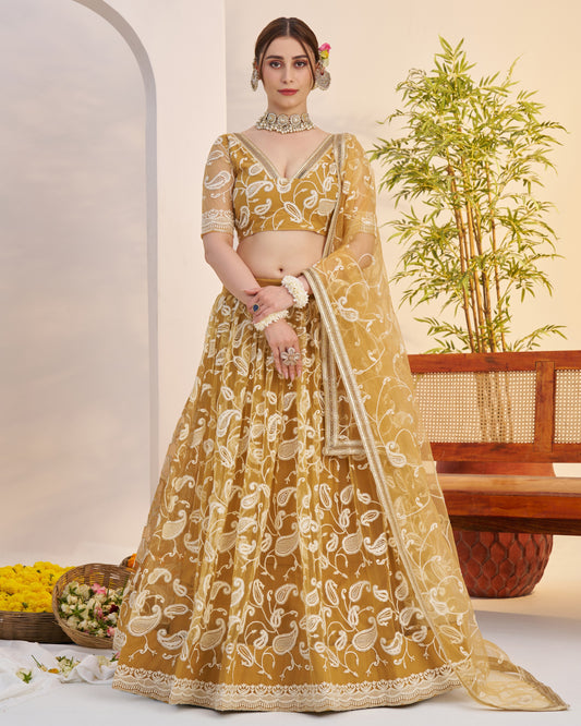 Woman in a mustard yellow traditional outfit with white patterns, standing indoors with a plant and wooden furniture in the background.