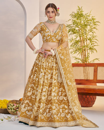 Woman in a yellow and white patterned traditional outfit standing indoors with a plant and basket in the background.
