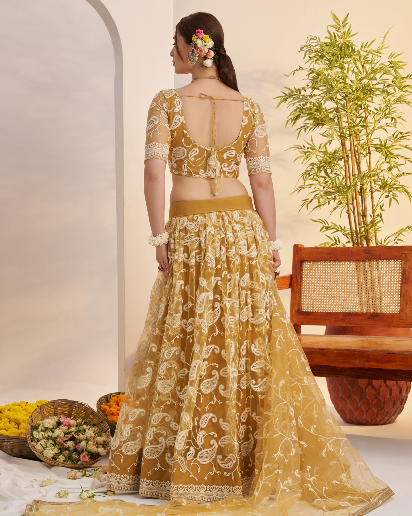 Woman in a yellow traditional outfit standing in a decorated indoor setting with flowers and a plant.