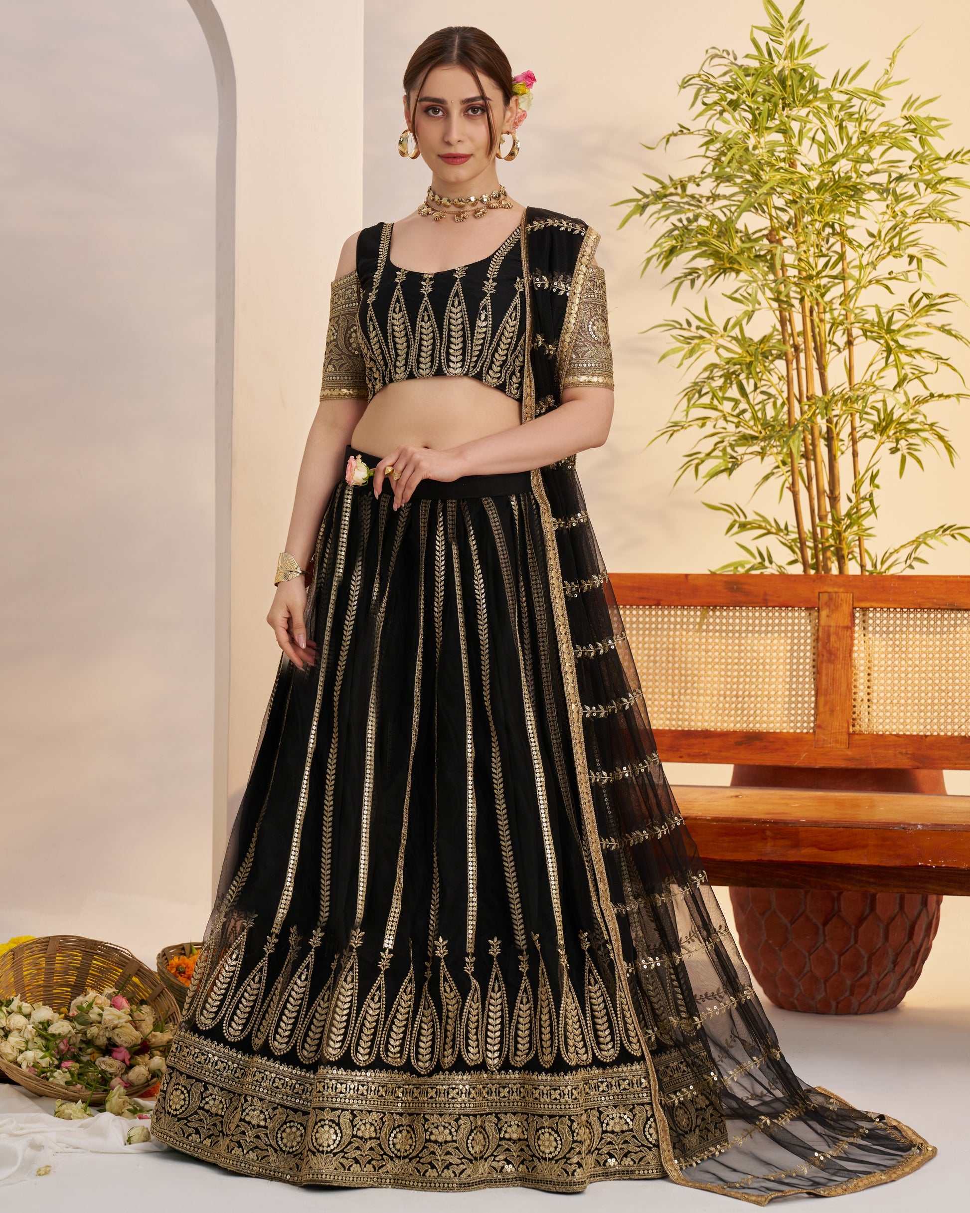 Woman wearing a black and gold embroidered traditional outfit with a plant and bench in the background.