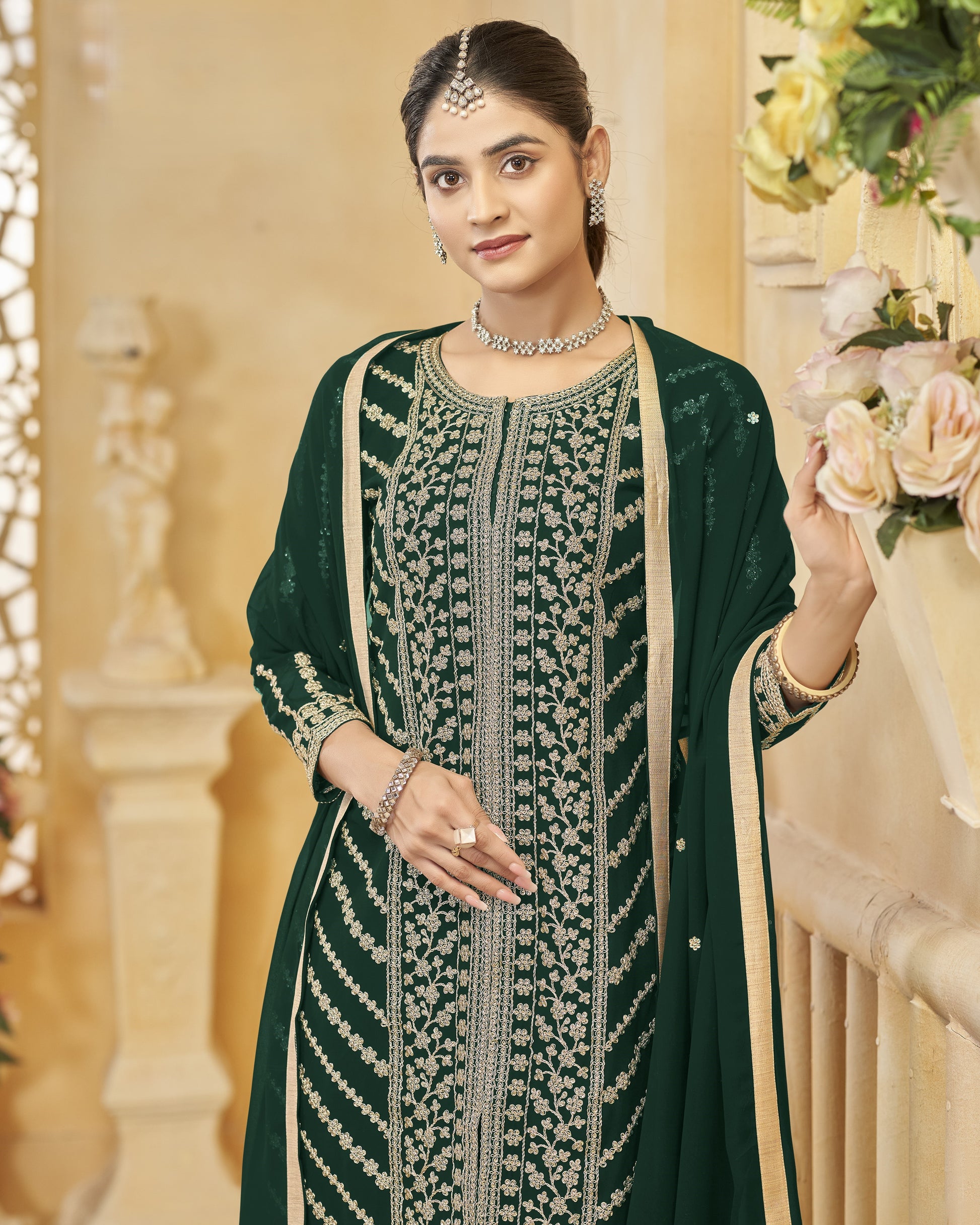 Woman in a green embroidered traditional outfit holding flowers indoors.