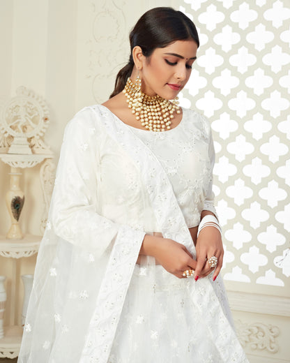 Woman in a white traditional outfit with gold jewelry against a decorative white wall.