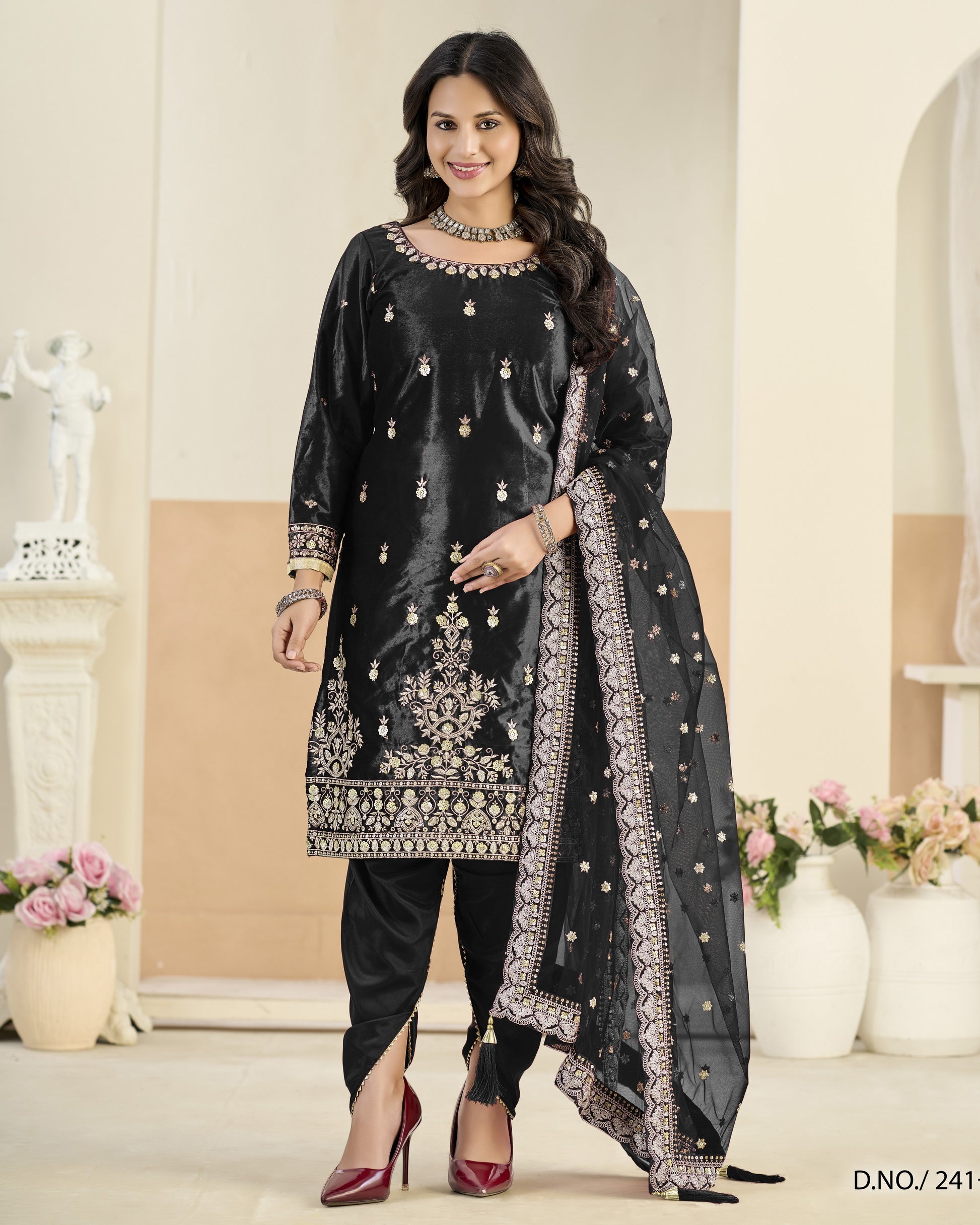 Woman wearing a black embroidered traditional outfit with a sheer dupatta in a decorated room.