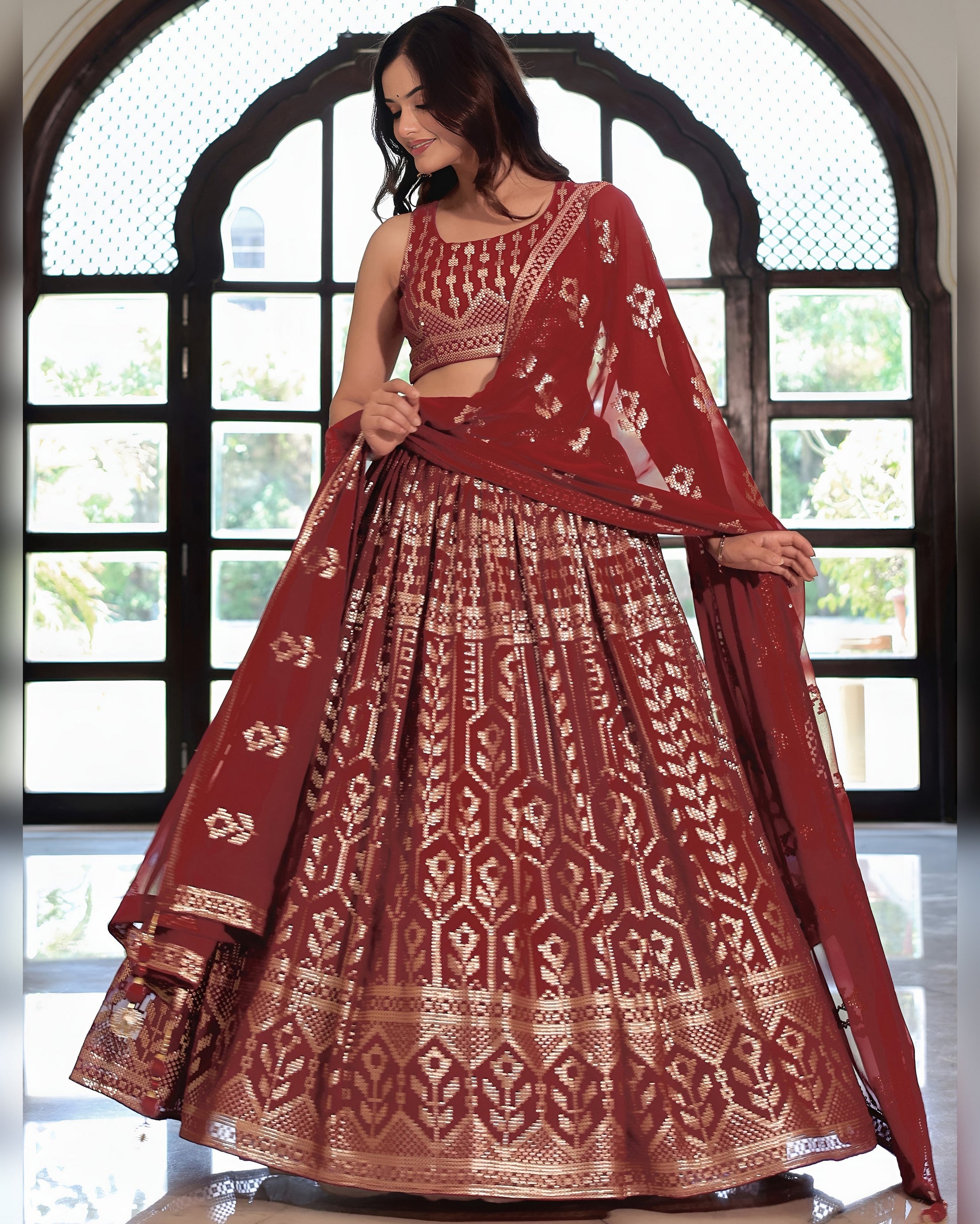Woman wearing a traditional maroon and gold embroidered outfit in front of a decorative window.