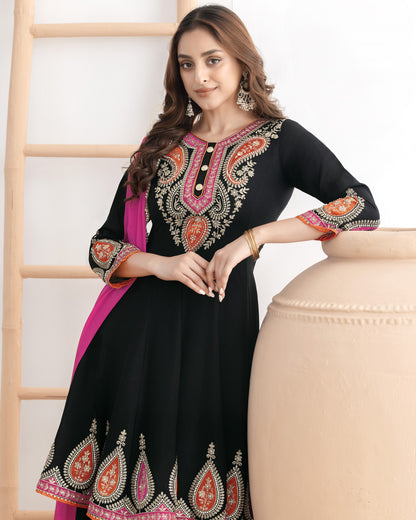 Woman wearing a black embroidered dress with colorful patterns, sitting on a beige chair.